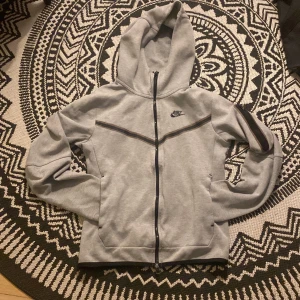 Grå hoodie från Nike - Säljer en grå hoodie från Nike med dragkedja och huva. Den har svarta detaljer och en liten Nike-logga på bröstet. Perfekt för en avslappnad stil. 
