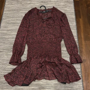 Röd och svart mönstrad klänning - Snygg röd och svart klänning med zebramönster. Klänningen har långa ärmar och en smockad midja som ger en fin passform. Perfekt för en trendig look!