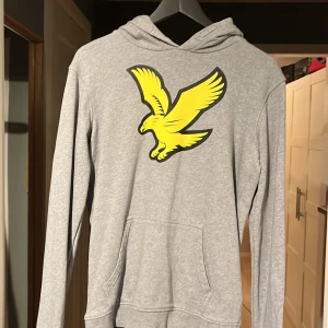 Grå hoodie från Lyle & Scott - Säljer en stilren grå hoodie från Lyle & Scott med en stor gul örn på bröstet. Tröjan har långa ärmar och en praktisk ficka framtill. Perfekt för en avslappnad look.
