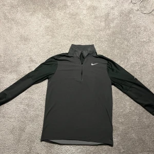 Svart långärmad tröja från Nike - Säljer en stilren svart långärmad tröja från Nike med dragkedja vid halsen. Perfekt för träning eller vardagsbruk. Tröjan har en enkel design med Nike-loggan på bröstet.