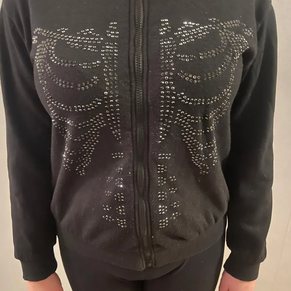 Snygg svart hoodie med dragkedja och paljettdetaljer i form av ett skelettmönster på framsidan. Perfekt för en cool och avslappnad stil. Den har långa ärmar och en bekväm passform.. Neuletakit & Villapaidat.