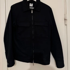 Svart jacka från Zara - Snygg mörkblåa jacka från Zara med dragkedja framtill och två bröstfickor med lock. Perfekt för en stilren look. Jackan har långa ärmar och en klassisk krage. Pris kan diskuteras, storlek M funkar även som S