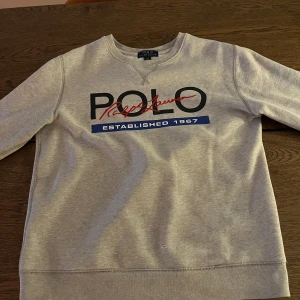 Grå tröja från Polo Ralph Lauren - Säljer en stilren grå tröja från Polo Ralph Lauren med tryck framtill. Tröjan har rund hals och långa ärmar, perfekt för en avslappnad look. Den har ett klassiskt tryck med texten 'Established 1967'.