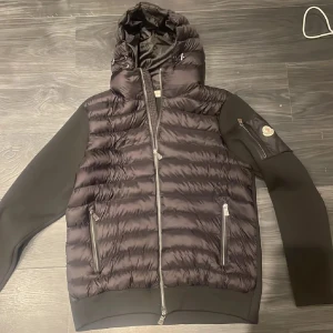 Moncler cardigan  - Säljer en snygg svart quiltad jacka från Moncler med luva och dragkedja. Jackan har långa ärmar och praktiska fickor med dragkedjor. Perfekt för kyligare dagar med stil. 🖤