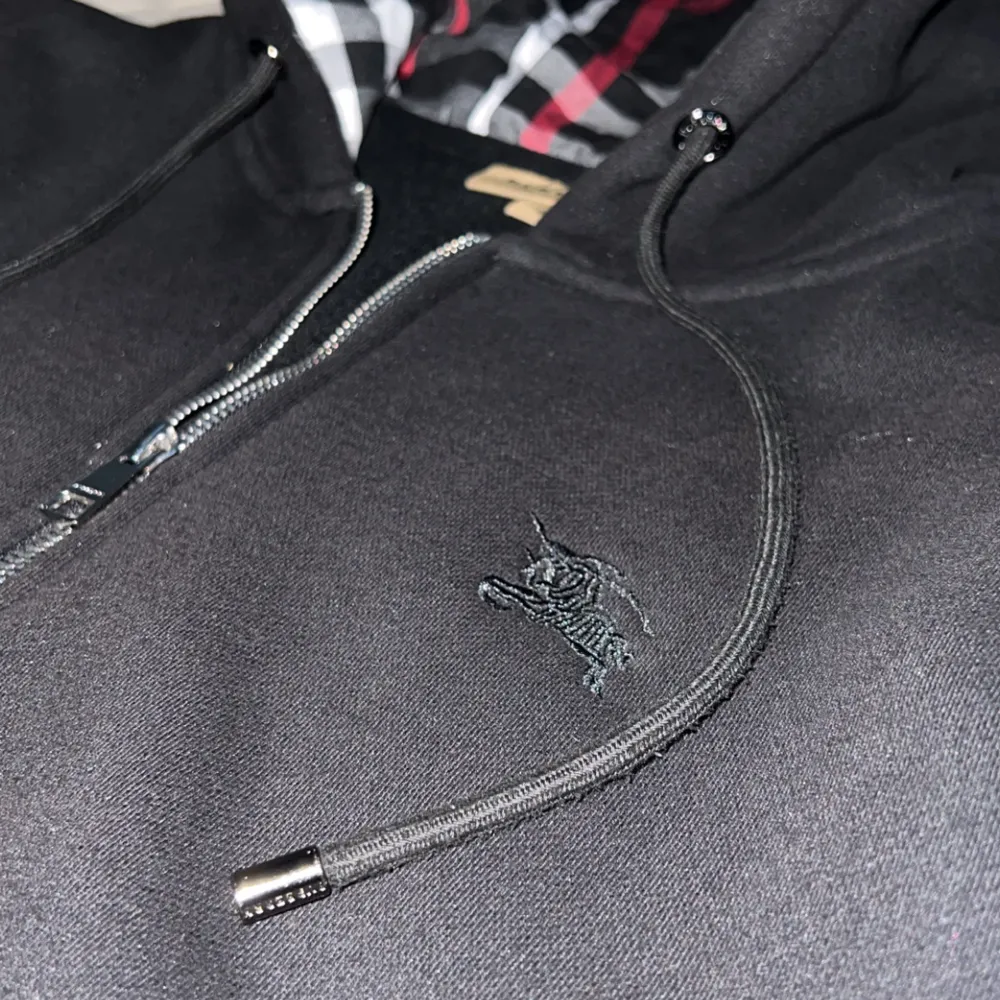 Säljer en stilren svart hoodie med dragkedja och en mönstrad insida i huvan. Tröjan har långa ärmar och praktiska fickor framtill. Perfekt för en avslappnad look.. Neuletakit & Villapaidat.