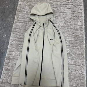 Beige hoodie och byxor från Boss - Säljer en stilren beige hoodie och matchande byxor från Boss. Hoodien har dragkedja och svarta ränder längs ärmarna. Byxorna har samma svarta ränder längs benen och en bekväm passform. Perfekt för en avslappnad look.ny köpt för 4200 kr mitt pris 2500 kr använd endast 7 gånger.kan ta byte mot en 14 pro max här i sundsvall bara ni får byte en iphone 14 pro 128gb batterihälsa 83% plus boss dressen mot 14 pro max.