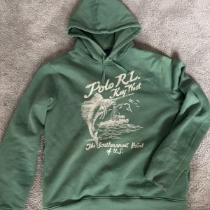 Grön hoodie från Polo Ralph Lauren - Säljer en snygg grön hoodie från Polo Ralph Lauren med tryck av en fisk och texten 'Key West' på framsidan. Perfekt för en avslappnad stil med en touch av äventyr. Huvan har dragsko och tröjan har en klassisk passform. Använt ett fåtal gånger 