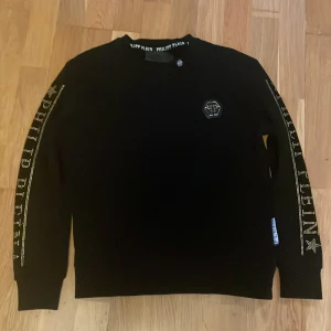 Svart tröja från Philipp Plein - Snygg svart tröja från Philipp Plein i bomull. Tröjan har långa ärmar med märkets namn i guld längs ärmarna och en cool logotypdetalj på bröstet. Perfekt för en stilren look. Skicka gärna en prisförslag om du vill minska det.