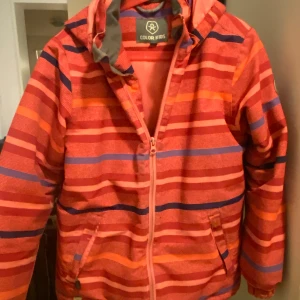 Färgglad randig jacka från Color Kids - Säljer en färgglad jacka från Color Kids i storlek 140/146. Jackan har ett randigt mönster i rött, orange och blått med dragkedja framtill och en praktisk huva. Det finns en reva i tyget baktill (se bild) annars är jackan i fint skick! 