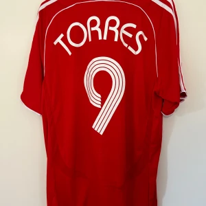 Röd Liverpool fotbollströja med Torres 9 - Säljer en röd Liverpool fotbollströja från Adidas med Torres och nummer 9 på ryggen. Tröjan har korta ärmar och klassisk design med Carlsberg-logga på framsidan. Perfekt för alla Liverpool-fans!