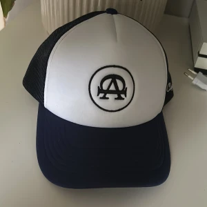 Vit och marinblå keps med nät - Säljer en snygg keps från All Caps Co. med vit framsida och marinblå skärm. Kepsen har ett svart broderat emblem på framsidan och text på sidan. Baksidan är i svart nätmaterial för bra ventilation. Perfekt för sommardagar!