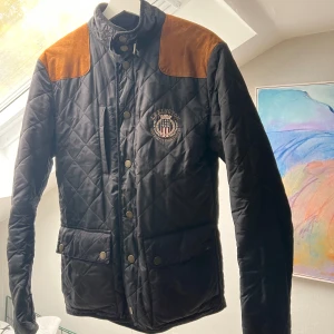 Svart quiltad jacka från Lexington - Säljer en snygg svart quiltad jacka från Lexington med bruna axelpartier. Jackan har knappar framtill och en cool logga på bröstet. Perfekt för höst och vår när du vill hålla stilen. Passar både till vardags och lite finare tillfällen. 