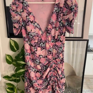 Blommig klänning med volangdetaljer -  blommig klänning i rosa och lila toner. Den har en v-ringad hals och korta puffärmar. Klänningen har volangdetaljer framtill och en åtsittande passform som framhäver figuren.