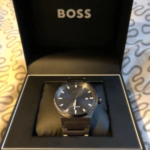 Svart automatisk klocka från BOSS - Säljer en stilren och elegant automatisk klocka från BOSS. Klockan har en svart urtavla med datumvisning och silverfärgade markeringar. Armbandet är i matchande svart metall och ger en modern look. Perfekt för både vardag och fest! Kvitto finns