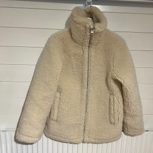 Beige teddyjacka från Arket - Säljer en supermysig beige teddyjacka från Arket. Den har en dragkedja framtill och är perfekt för kyliga höst- och vinterdagar. Jackan är långärmad och har en normal till oversized passfotm. Den är i nyskick och redo att hålla dig varm och stilren!