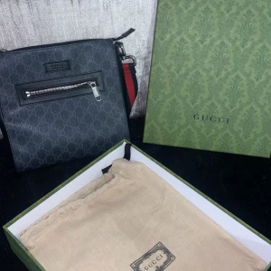 Gucci messenger  - Snygg svart axelväska från Gucci med ett diskret mönster och en praktisk dragkedja framtill. Väskan har en justerbar axelrem i rött och grönt, vilket ger en klassisk touch. Perfekt för att bära dina nödvändigheter med stil.