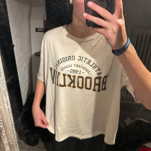 Vit t-shirt från Gina Tricot  - Säljer en gräddvit t-shirt från Noisy May med trycket 'Brooklyn Athletic Original 1995' i svart och gult. T-shirten har en loose/oversize passform och korta ärmar, perfekt för en avslappnad stil.