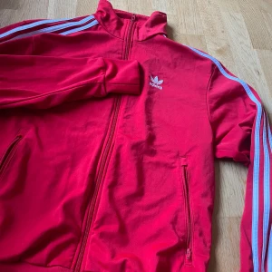 Röd träningsjacka från Adidas - Snygg röd träningsjacka från Adidas med de klassiska tre ränderna i vitt längs ärmarna. Jackan har dragkedja framtill och en hög krage. Perfekt för en sportig look.
