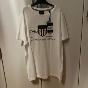 Vit t-shirt från GANT - Snygg vit t-shirt från GANT med tryck av deras klassiska logga och texten 'American Sportswear'. Perfekt för en avslappnad stil. Denna tröja är även helt ny med pris lapp på.