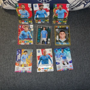Fotbollskort från Manchester City - Samling av fotbollskort med spelare från Manchester City. Inkluderar kort med etiketter som 'Rising Stars', 'Playmaker', 'International Star' och 'Future Star'. Kortens design har färgglada bakgrunder och visar spelare i lagets blå matchtröjor. Perfekt för samlare och fotbollsfans.vill ha mer bilder bara fråga