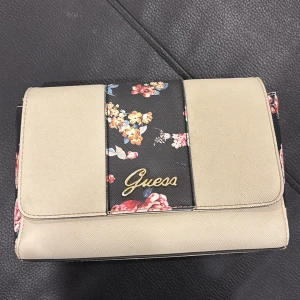 Beige och blommig axelväska från Guess - Snygg beige axelväska från Guess med blommigt mönster och guldfärgad kedja. Väskan har en elegant design med Guess-logga på framsidan och justerbar axelrem. Perfekt för att ge en färgklick till din outfit! Original pris 1300kr. Använd en gång, säljer för den inte passar min stil 