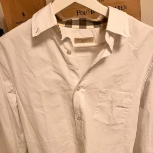 Vit skjorta från Burberry - Snygg vit skjorta från Burberry med klassisk krage och knappstängning. Skjortan har en bröstficka med diskret broderad logga och ett rutigt mönster på insidan av kragen. Perfekt för en stilren look.