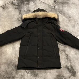 Svart dunjacka med pälsdetalj - Säljer en svart dunjacka från Canada Goose med avtagbar huva och pälsdetalj. Jackan har långa ärmar och stängs med dragkedja och knappar. Perfekt för kalla vinterdagar. Det är bara två knappar som inte fungerar man ser det på sista bilden.