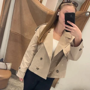 Beige trenchcoat från Vero Moda - Säljer denna jättefina beigea trenchcoat från Vero Moda då den inte kommit till användning många gånger. Jackan är croppad och stängs med knappar i fram. 