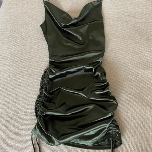 Mörkgrön satin klänning - Snygg mörkgrön klänning i satin med en smickrande drapering och snörning på sidorna. Klänningen har en v-ringad design och är ärmlös, vilket ger en elegant look.