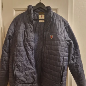 Fjällräven Dunjacka/Expedition - Säljer en blå quiltad jacka från Fjällräven, inspirerad av deras 1974 Expedition. Jackan har en dragkedja framtill och är långärmad med ett klassiskt Fjällräven-märke på bröstet. Perfekt för kyliga dagar.