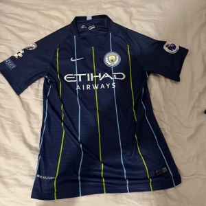 Blå Manchester City fotbollströja - Säljer en blå Manchester City fotbollströja från Nike med korta ärmar. Tröjan har gula och ljusblå ränder samt sponsorlogga på framsidan. På baksidan finns namnet 'De Bruyne' och nummer 17. Perfekt för fans av laget! Pris kan diskuteras