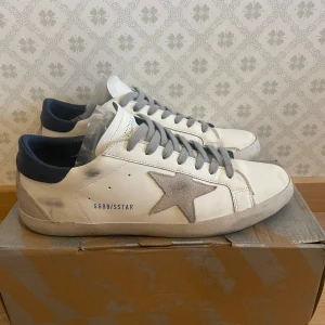 Vita sneakers från Golden Goose - Snygga vita sneakers från Golden Goose med grå stjärndetalj på sidan och mörkblå häl. Skorna har grå snörning och kommer med originalkartong och påse. Perfekta för en stilren look.