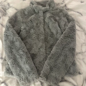 Grå teddyjacka från Vero Moda - Mysig grå teddyjacka från Vero Moda. Perfekt för kyliga dagar med sin fluffiga och mjuka textur. Jackan har långa ärmar och en stilren design som passar till många olika outfits. Kontakta mig om ni har frågor.
