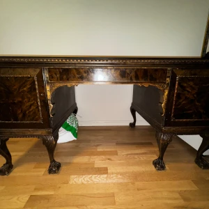 Antikt skrivbord i mörkt trä - Elegant antikt skrivbord i mörkt trä med detaljerade sniderier och lejonfötter. Skrivbordet har en central låda och skåp på sidorna med vackra fanérdetaljer. Perfekt för ett klassiskt inrett kontor eller vardagsrum. 