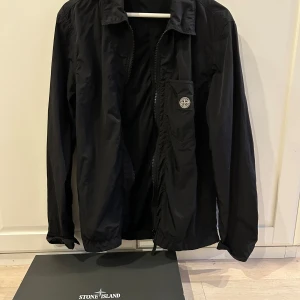 Svart jacka/overshirt från Stone Island - Säljer en stilren svart overshirt från Stone Island med dragkedja och klassisk krage. Jackan har en diskret bröstficka med det ikoniska Stone Island-märket. Perfekt för lager-på-lager-stil.