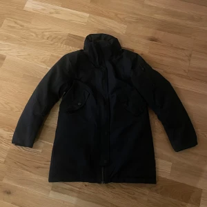 Svart parkajacka med dragkedja - Snygg svart parkajacka med dragkedja och knappar framtill. Jackan har långa ärmar och praktiska fickor på bröstet. Perfekt för kyligare dagar.