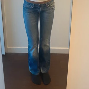 Low waist jeans - Säljer ett par blå bootcut jeans med slitna detaljer och klassisk femficksdesign. Jeansen har en låg midja och är perfekta för en avslappnad stil. De har en snygg tvättad look och är tillverkade i denim.