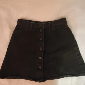 Svart jeanskjol från Monki - Snygg svart jeanskjol från Monki i storlek 38. Kjolen har knappar framtill och två bakfickor. Perfekt för en cool och avslappnad stil.