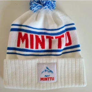 Vit mössa med Minttu-logga - Snygg vit mössa med blå och röda detaljer från Minttu. Mössan har en ribbad kant och en blå-vit tofs på toppen. Perfekt för kalla dagar!