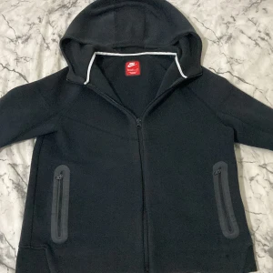 Nike tech fleece - Säljer en svart fleecejacka från Nike med huva och dragkedja. Koftan har praktiska fickor med dragkedjor och är perfekt för kyligare dagar. Den är långärmad och har en stilren design.
