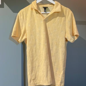 Gul froté-tröja från H&M - Säljer en gul pikétröja från H&M med ett subtilt palmmönster. Tröjan har korta ärmar och en klassisk krage. Perfekt för sommarn som kommer.  Tröjan är använd en gång. Nypris är 500kr. Mitt pris 350.