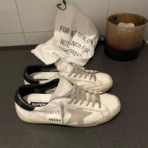 Golden Goose Super-Star sneakers - Säljer ett par Golden Goose Super-Star sneakers i vitt med grå detaljer och svart häl. Skorna har en ikonisk stjärndetalj på sidan och snörning framtill. Perfekta för en avslappnad stil.