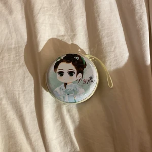 Rund plånbok med tecknad design - Söt rund plånbok med en tecknad karaktär på framsidan. Den har en dragkedja runtom och en liten rem för enkel bärning. Perfekt för att förvara småsaker eller mynt. 💕