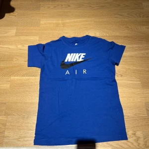 Blå Nike T-shirt med logga - Säljer en blå Nike T-shirt med den klassiska Nike Air-loggan i vitt och svart på framsidan. Perfekt för en sportig look eller avslappnad stil. Tillverkad i mjukt material för extra komfort.