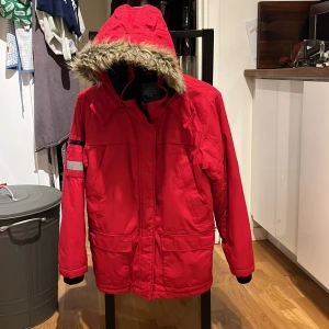 Röd vinterjacka från Lindex - Säljer en röd vinterjacka från Lindex med avtagbar huva och fuskpäls. Jackan har långa ärmar och flera praktiska fickor. Perfekt för kalla dagar.