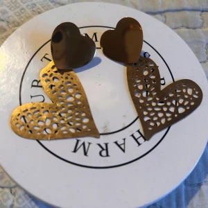 Guldiga hjärtformade örhängen - Charmiga örhängen i guld med hjärtformad design. De har en solid hjärtformad topp och ett större, perforerat hjärta som hänger nedanför. Perfekta för att ge en romantisk touch till din stil.