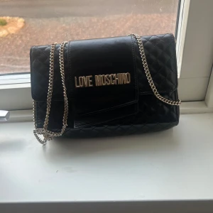 Svart quiltad axelväska från Love Moschino - Snygg svart axelväska från Love Moschino med quiltad design och kedjeaxelrem. Väskan har en elegant logotypdetalj på framsidan och är perfekt för att ge din outfit en stilfull touch.