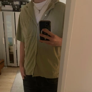Grön kortärmad frottéskjorta från H&M - Säljer en grön kortärmad skjorta från H&M i regular fit. Skjortan har en bröstficka och knappar framtill. Sparsamt använd. Perfekt till ett par linnebyxor till sommaren