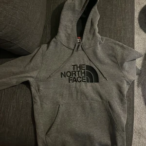 Grå hoodie från The North Face - Säljer en grå hoodie från The North Face med svart logga på bröstet. Tröjan har en klassisk design med huva och dragsko samt en stor magficka framtill. Perfekt för kyliga dagar.