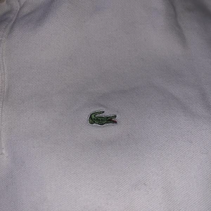 Vit pikétröja från Lacoste - Säljer en klassisk vit pikétröja från Lacoste med den ikoniska krokodilloggan på bröstet. Tröjan har korta ärmar och en traditionell krage med knappar. Perfekt för en stilren och avslappnad look.pris kan förhandlas 